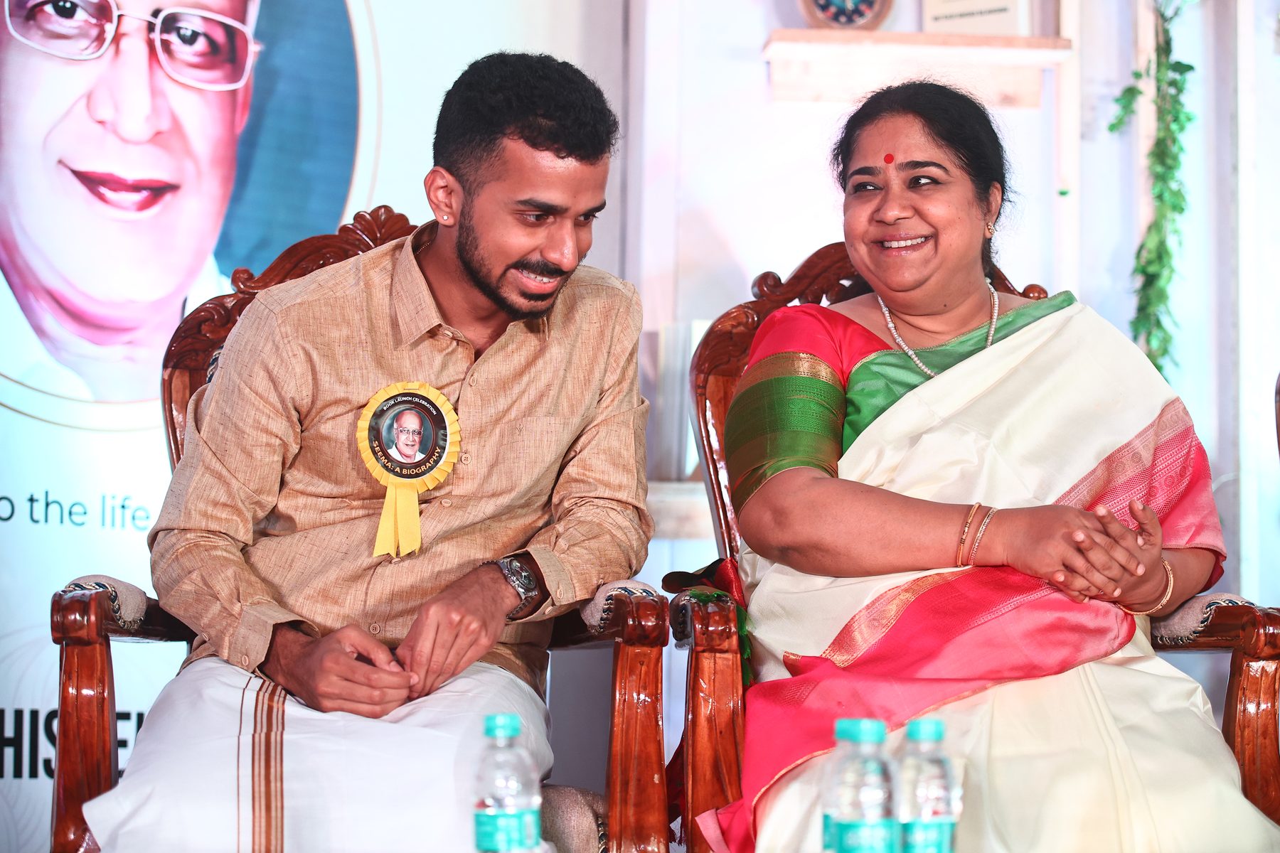 Achi Bala and Smt. Lalgudi Vijayalakshmi sharing a smile on stage at the Seema 90 book launch, seated before a banner of his grandfather