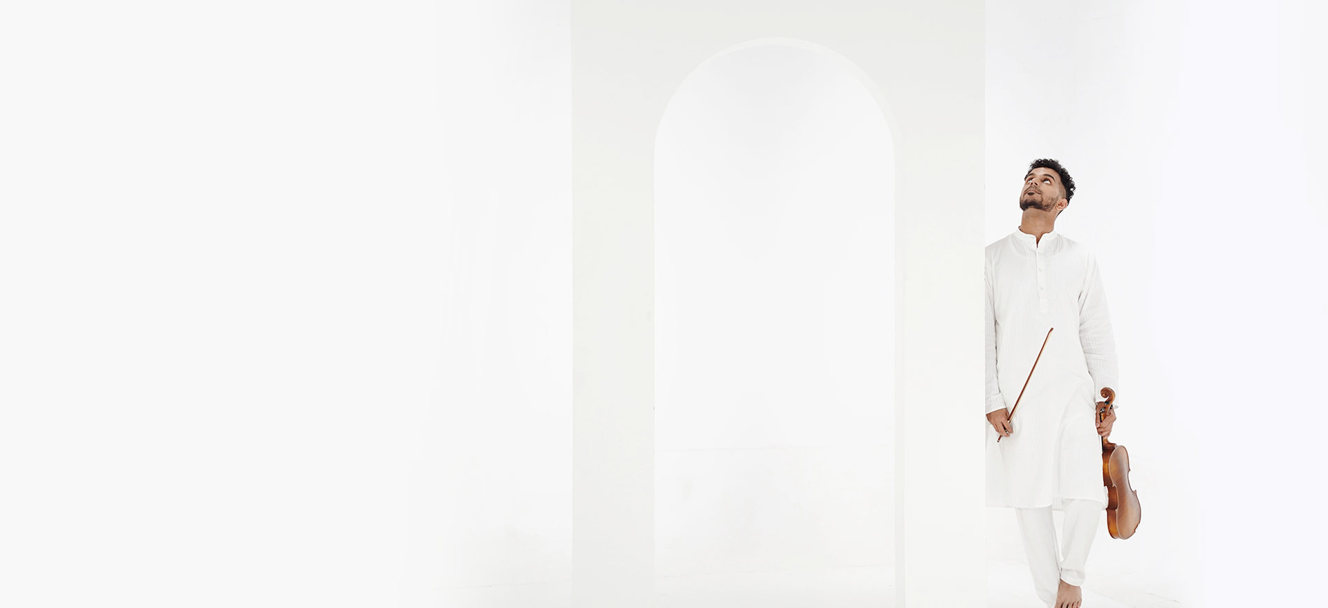 Achi Bala looking upward, holding his violin, in a bright white archway